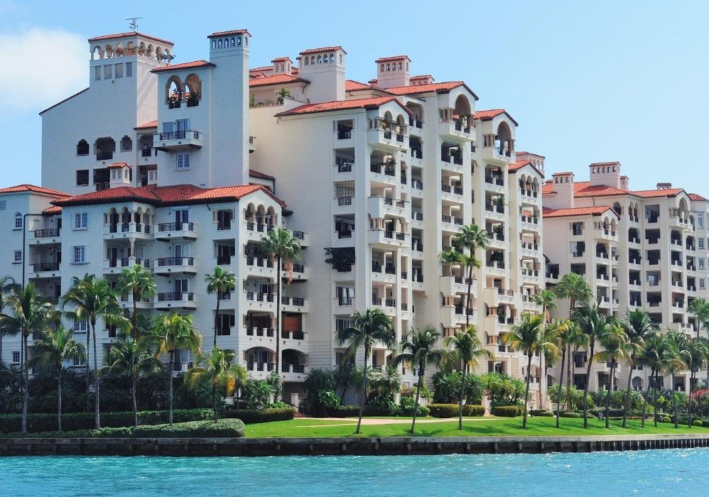 An image of a large apartment complex right on the ocean. Palm trees line the green space between the apartments and the ocean. 