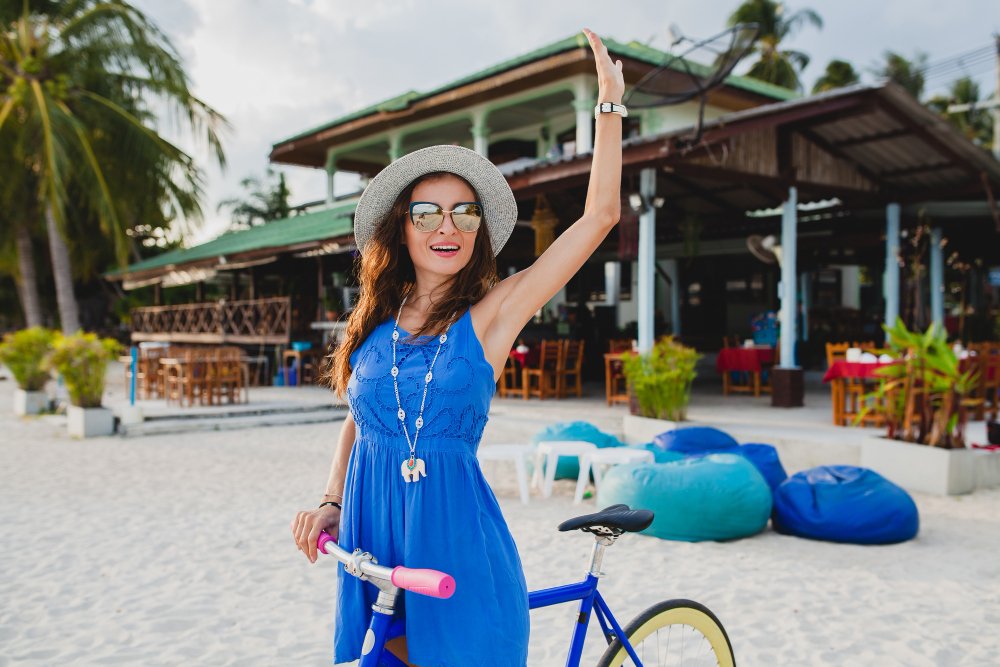 An image of a woman in a bright blue dress who has just rode her bike to the beach. She's waiving down her friends to enjoy dinner at a restaurant on the beach. 